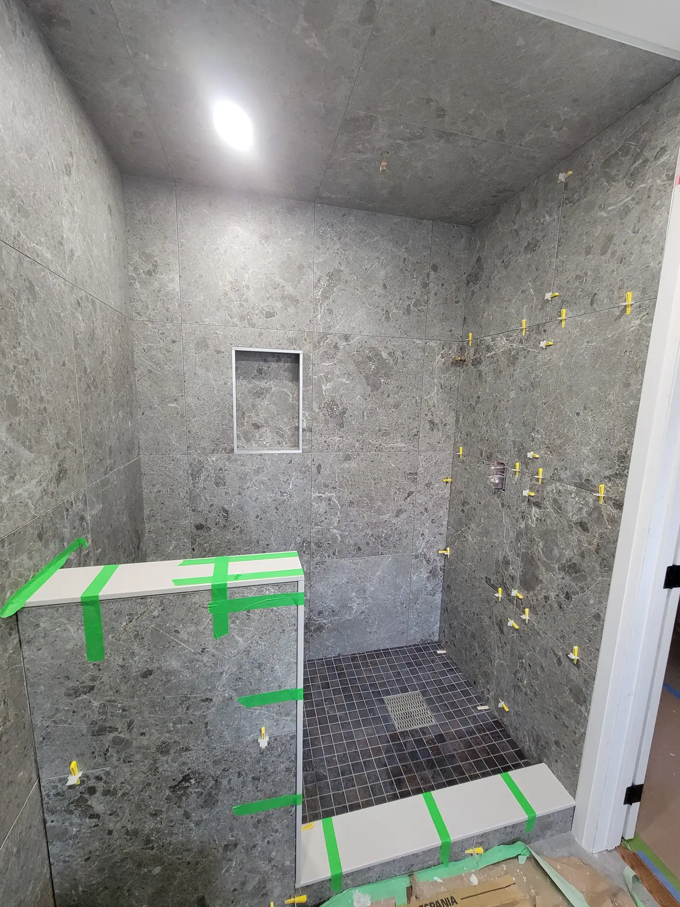 Large-format grey marble-look porcelain being set with levelling clips and a dark mosaic accent band in the Richmond Hill shower