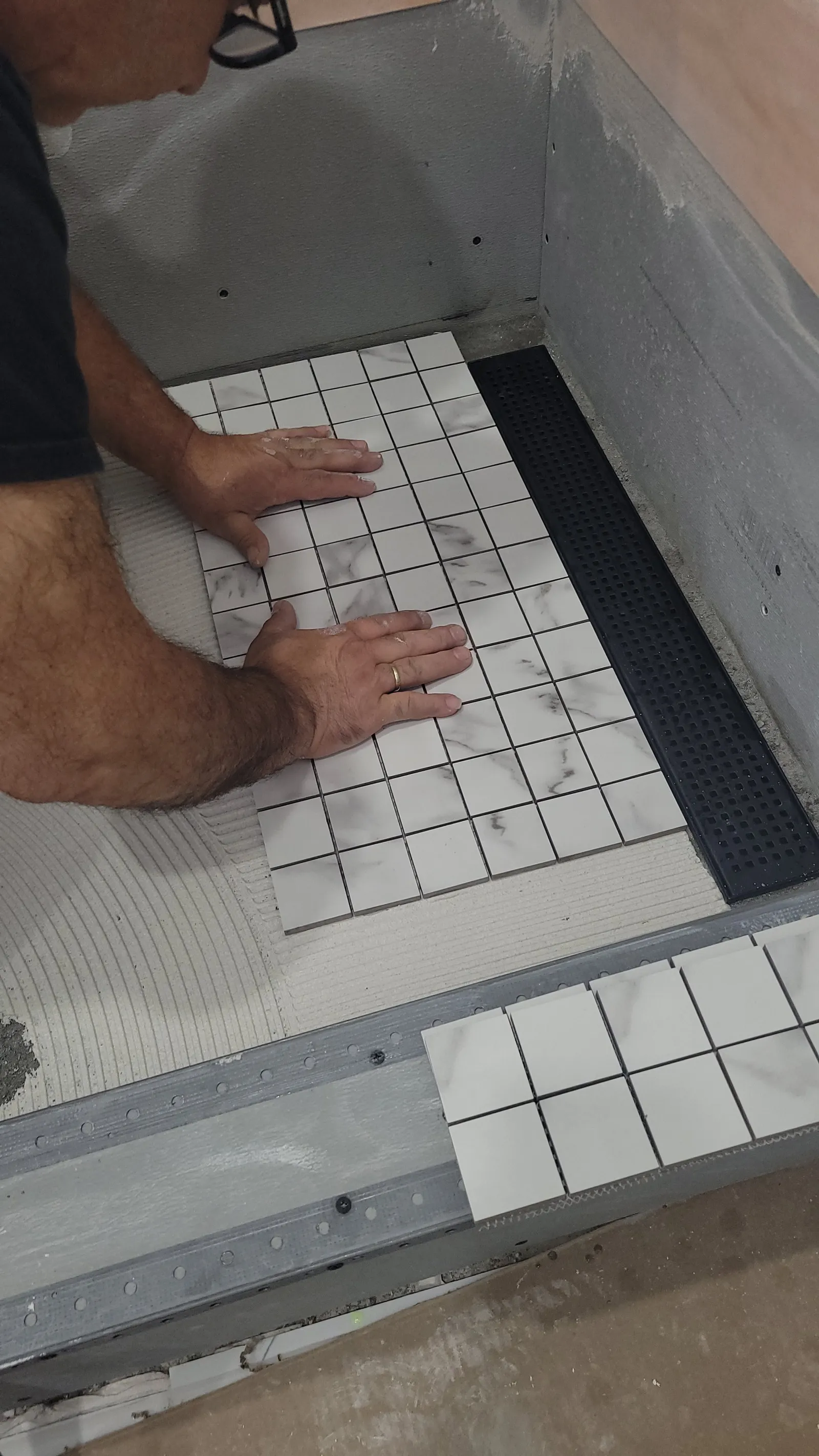 Hands pressing a sheet of marble-look porcelain mosaic onto the new shower floor beside the black linear drain in Newmarket