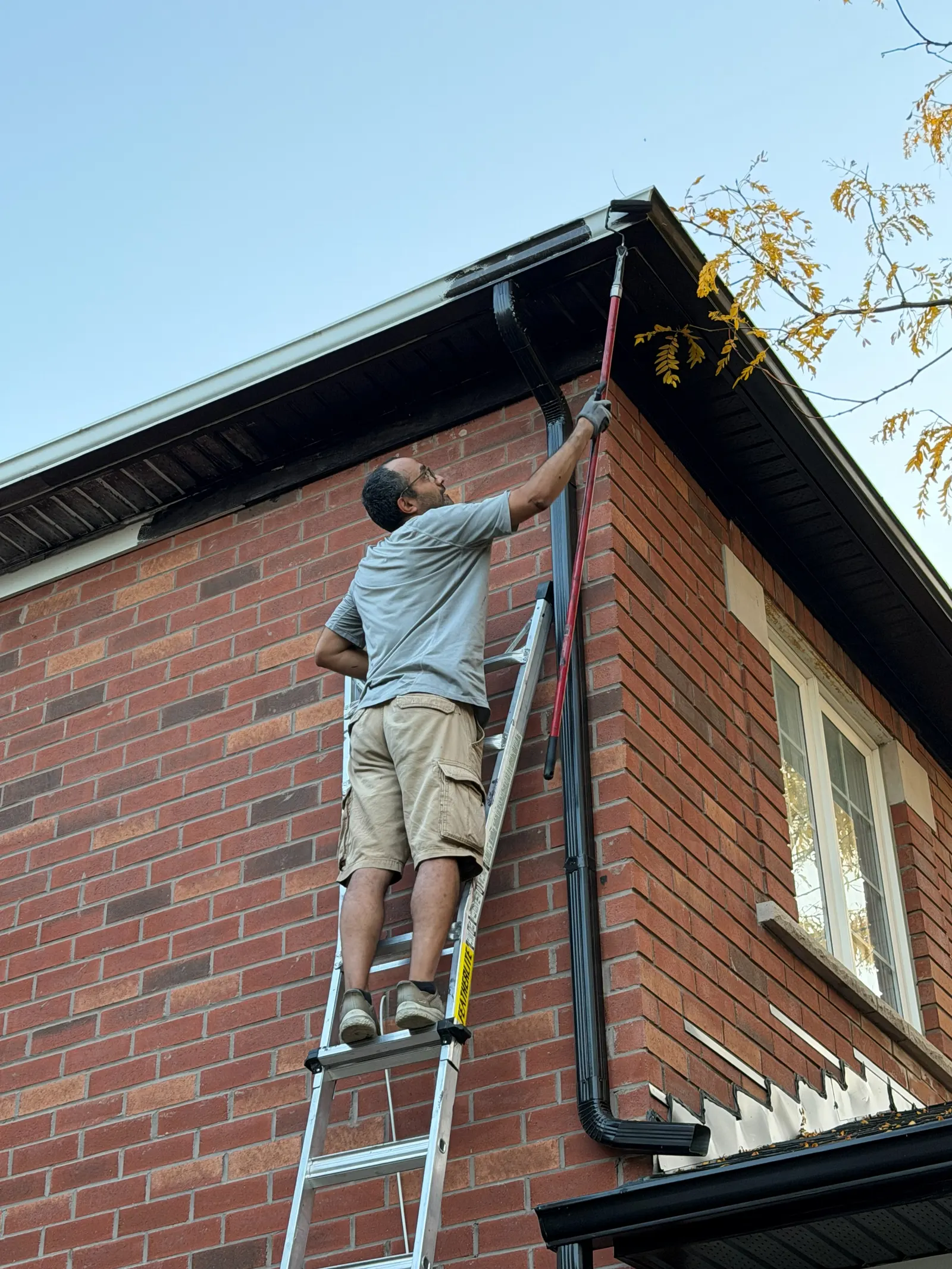 VOL Handyman team member on an extension ladder painting the dark soffit of a two-storey brick home with a roller on a red extension pole